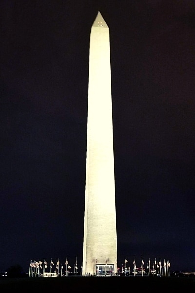 Washington Monument obelisk surrounded by a circle of American flags and lit up for the evening against a black sky.