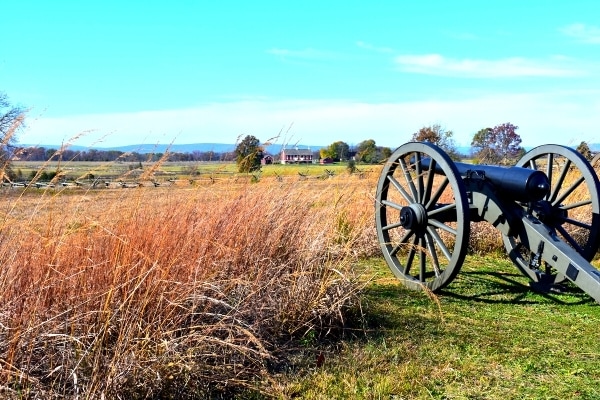 A cannon peers through tall yellow grass across Gettysburg Battlefield under a blue sky