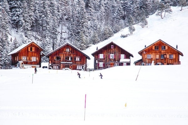 Skiers cruise through the thick snow in front of 4 wooden ski chalets in the snow covered woods of Malbun, Liechtenstein