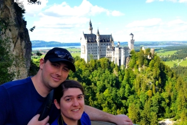 A couple smile for the camera with Neuschwanstein Castle, a green forest, and blue skies in the background