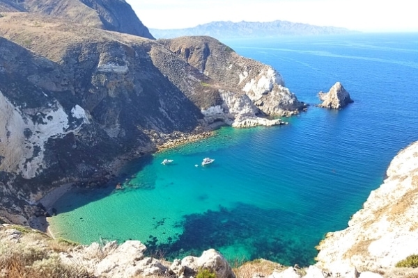 Tall cliffs of Santa Cruz Island surround Potato Harbor where the blue waters of the Santa Barbara Channel gradually turn aqua and then green as a small white sand beach appears against the bottom of the cliffs. Two small white boats float at anchor in the harbor.