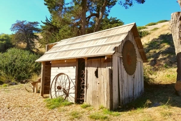 Simple wooden shed with metal wagon wheel leaning against it, surrounded by hills of Santa Cruz Island