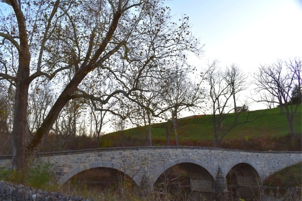 A three span stone arch bridge crosses Antietam Creek and is surrounded by tall hills