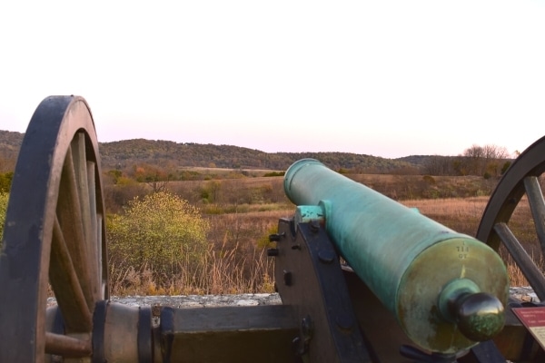 Looking down the side of a green-patinaed cannon aimed over the brown hillside in the distance