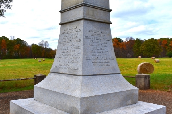 Base of a white stone grave marker in front of a farm field with hay bales and fall-colored trees in the background