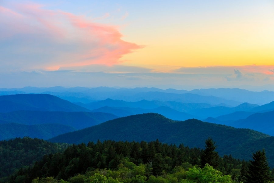 Layers on layers of blue mountain ridges lie under an orange sky as the sun sets at Cowee Mountain Overlook on Blue Ridge Parkway