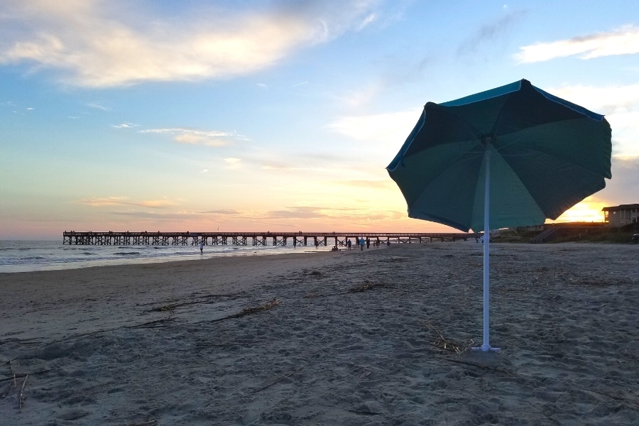 A blue and teal umbrella stands alone in the sand at Isle of Palms beach as the sun sets behind the pier in the distance