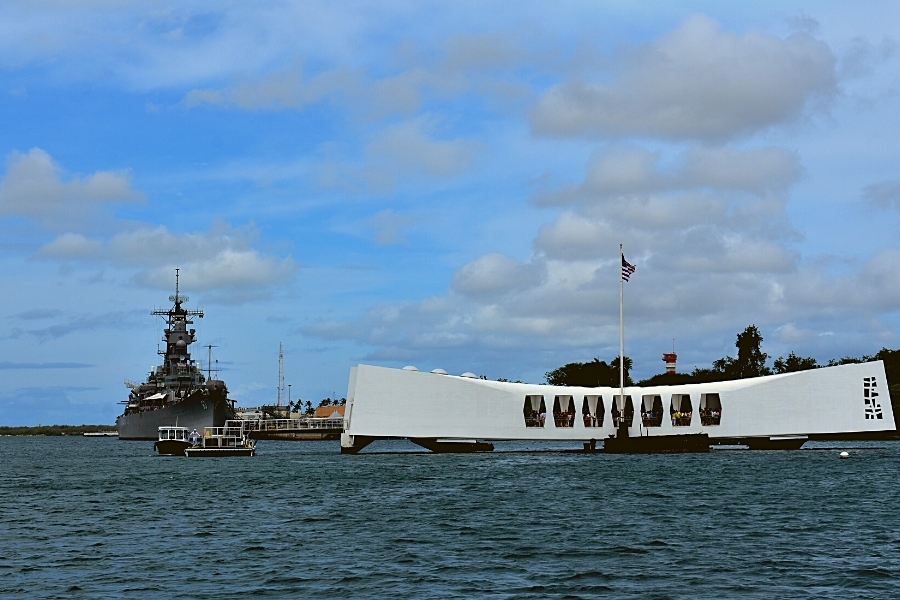 An American flag flies above the white USS Arizona Memorial in Pearl Harbor with the USS Missouri docked in the background.
