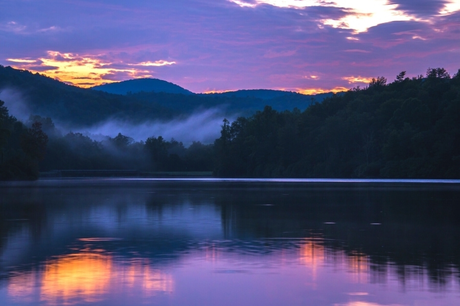 The cloudy sky turns purple as the sun rises behind the mountains surrounding Price Lake on Blue Ridge Parkway