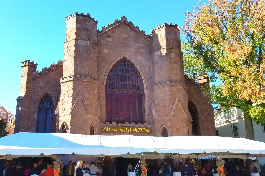 Large tan stone building with crenellated roofline and large cathedral windows houses the Salem Witch Museum. A white tarp out front protects visitors from the elements.