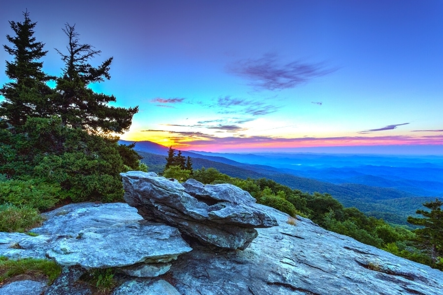 An orange sun rises behind the mountains, turning the dark blue sky orange, red, and purple over the Blue Ridge Mountains as seen from a rocky outcropping