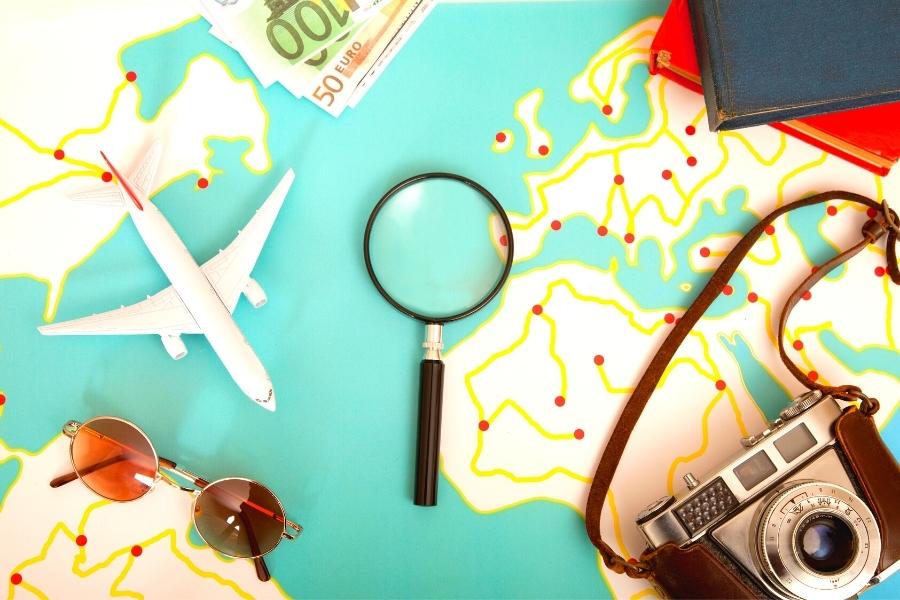Travel items like a camera, airplane, sunglasses, euros, and a magnifying glass, scattered over a world map table
