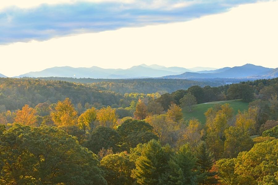 Orange, yellow, and green trees shine with the last light on an autumn day, looking out over the Biltmore Estate with the blue outlines of mountains on the horizon