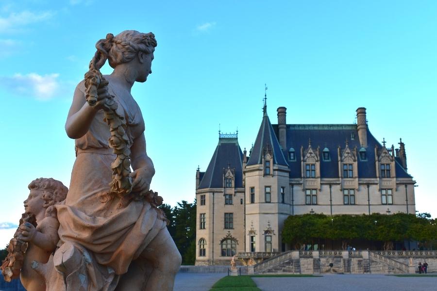 A white stone statue of a woman holding a floral rope looks back at the south terrace of the Biltmore House as the sun sets