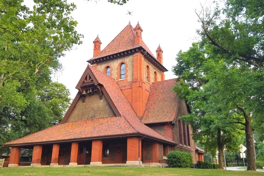 Large, multi-story red brick and red tile roofed church surrounded by green trees, The Cathedral of All Souls resides in Biltmore Village outside of the estate