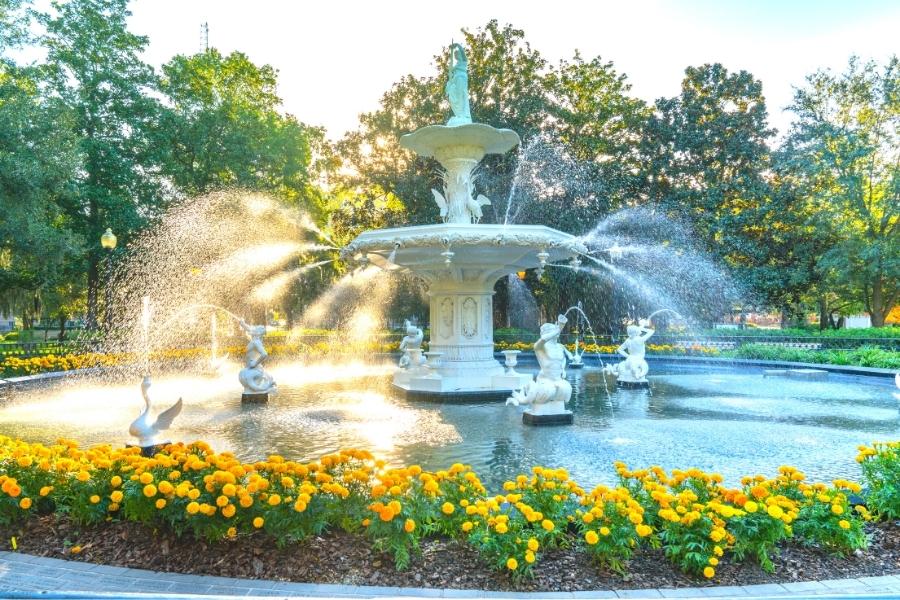 A two-tier, circular water fountain is surrounded by marigolds and green trees in Savannah, GA