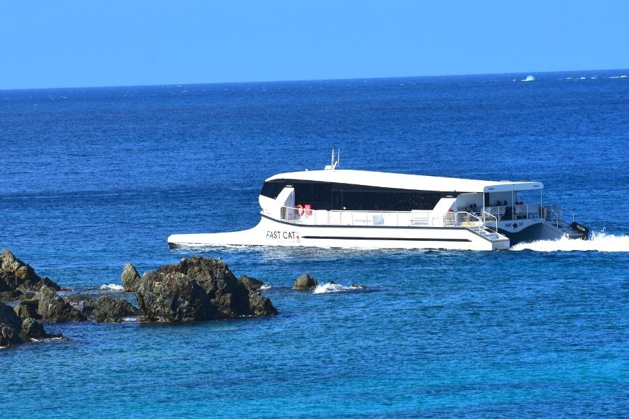 White, enclosed "Fast Cat" catamaran shuttle glides past St John's rocky shore on a blue ocean under a blue sky.