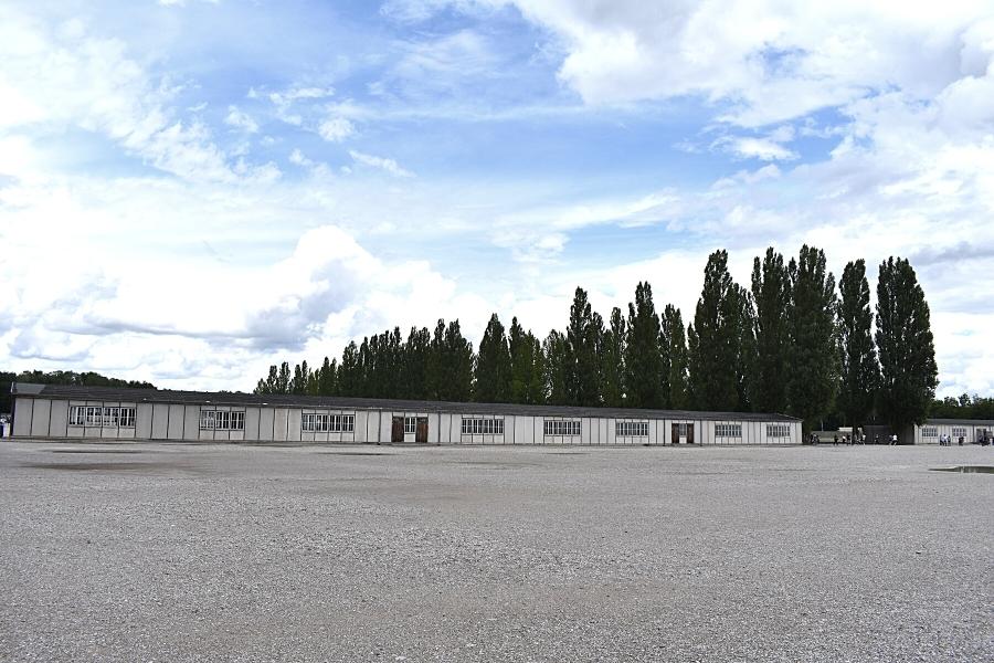 A large empty gravel roll-call yard sits empty with reconstructed gray barracks in the background