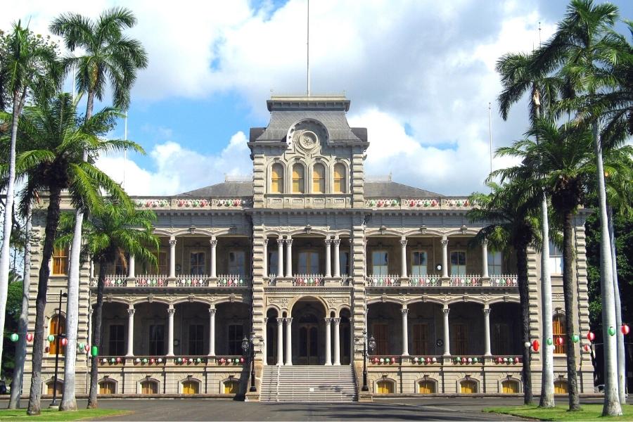 An imposing palace, once home to the Hawaiian monarchy, sits between an allee of palm trees under a blue cloudy sky
