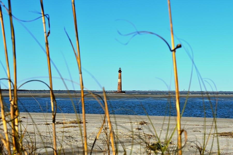 A brick lighthouse stands tall on a small island surrounded by the blue ocean and blue sky, with an empty beach and dune grass in the foreground