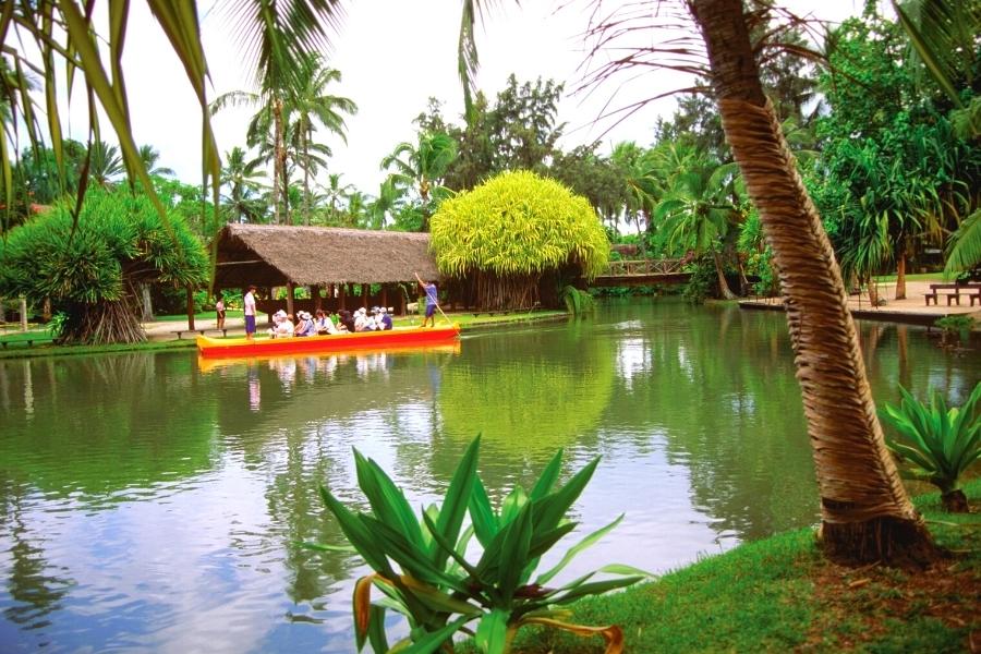 A canoe with tourists floats down the river surrounded by greenery and a thatched roof hut at the Polynesian Cultural Center in Hawaii