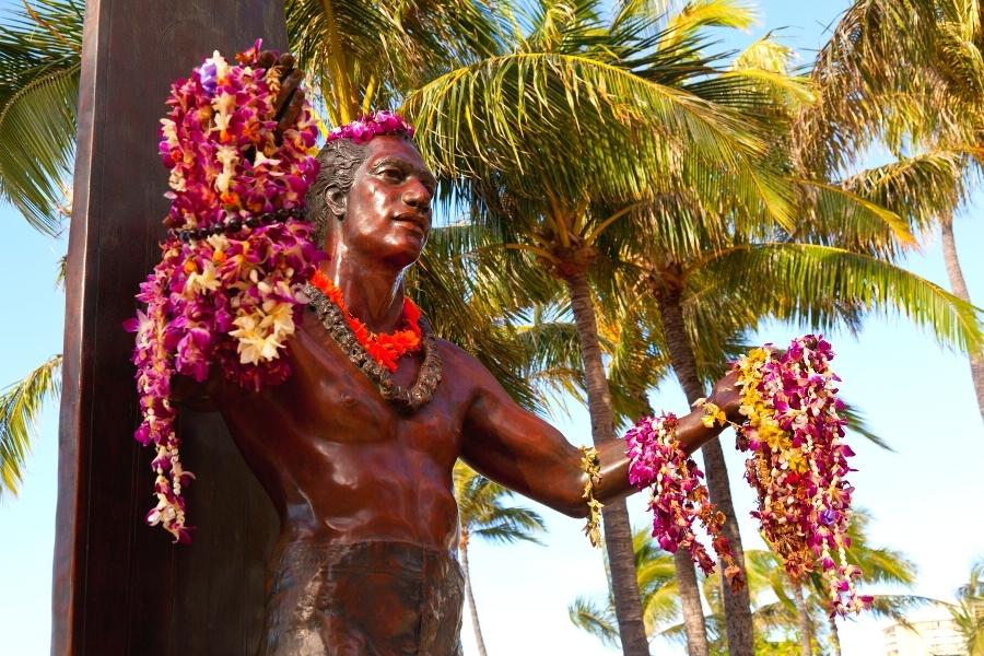 Bronze statue of Duke Kahanamoku at Waikiki Beach is surrounded by palm trees and draped with flower leis