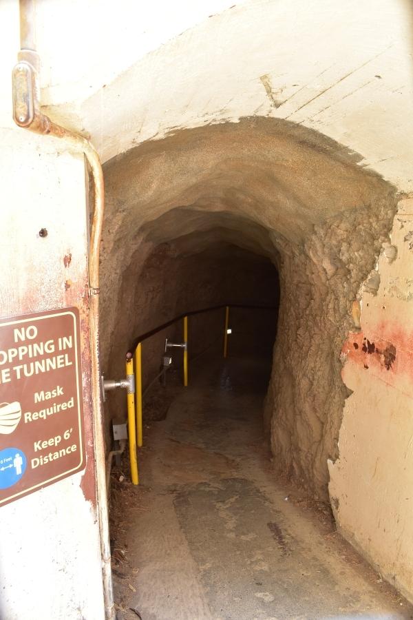 Dark tunnel leads into a craterside with yellow handrail and a sign requiring masks and social distancing and no stopping the tunnel