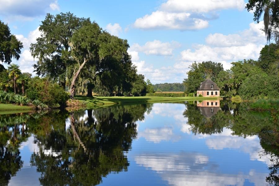 The Middleton Place Mill House is surrounded by green trees and blue sky, all reflected in the pond