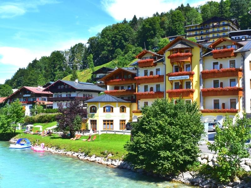 Colorful buildings with balcony flower boxes line the green hillside next to the blue river in Berchtesgaden, Germany