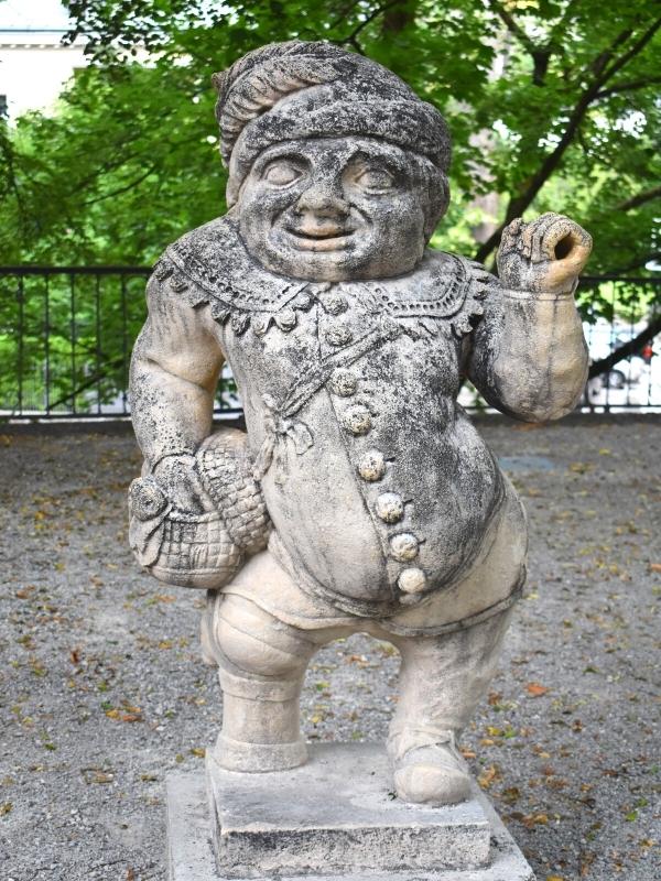 A small dwarf statue with a man's face, button down shirt, and a basket on his hip