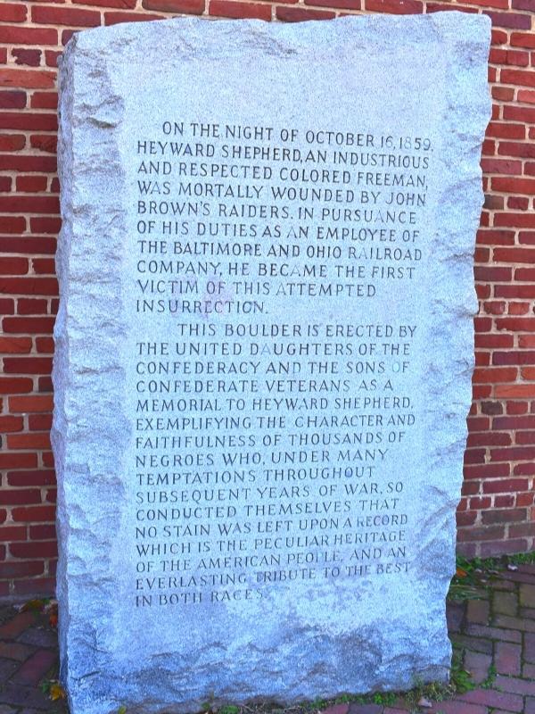 A simple granite memorial sits in front of a red brick building, inscribed with words memorializing Heyward Shepherd, in Harpers Ferry, WV