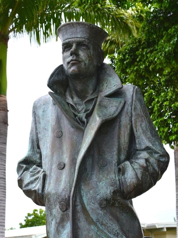 The Lone Sailor dark metal statue sits under palm trees and looks out at sea at the Pearl Harbor Memorial Site