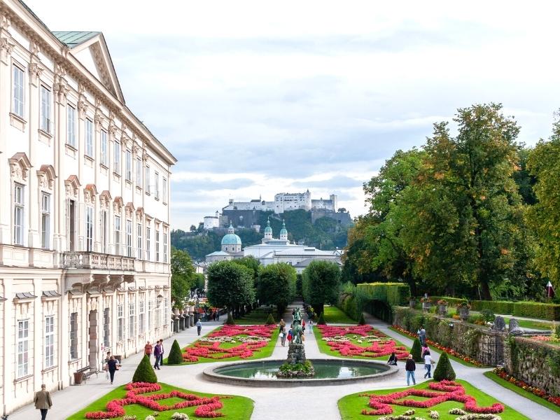 View looking down Mirabell Palace Gardens with the palace on the left, the gardens and fountains in the foreground, and Hohensalzburg Fortress in the background against a gray sky