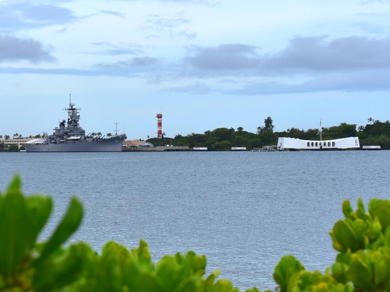 Ford Island as seen from the mainland of Oahu, with the white USS Arizona memorial and the gray USS Missouri battleship in the foreground and the orange and white tower at the Pearl Harbor Aviation Museum in the background