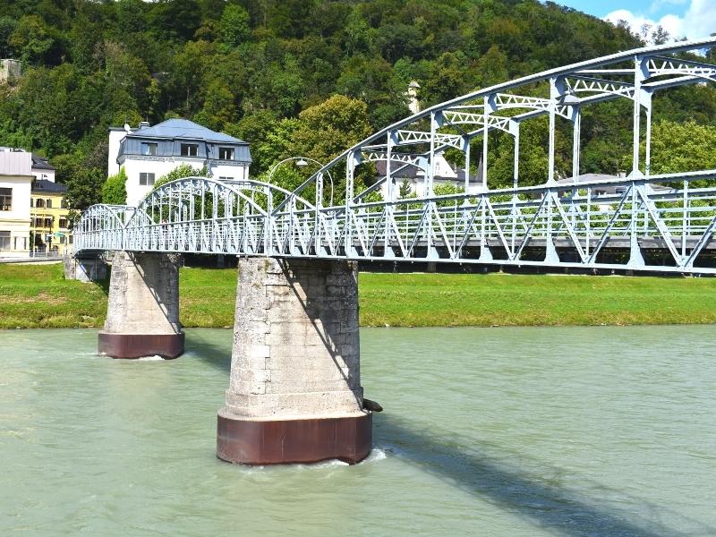 A blue metal arch pedestrian bridge crosses the blue Salzach River in Salzburg, Austria