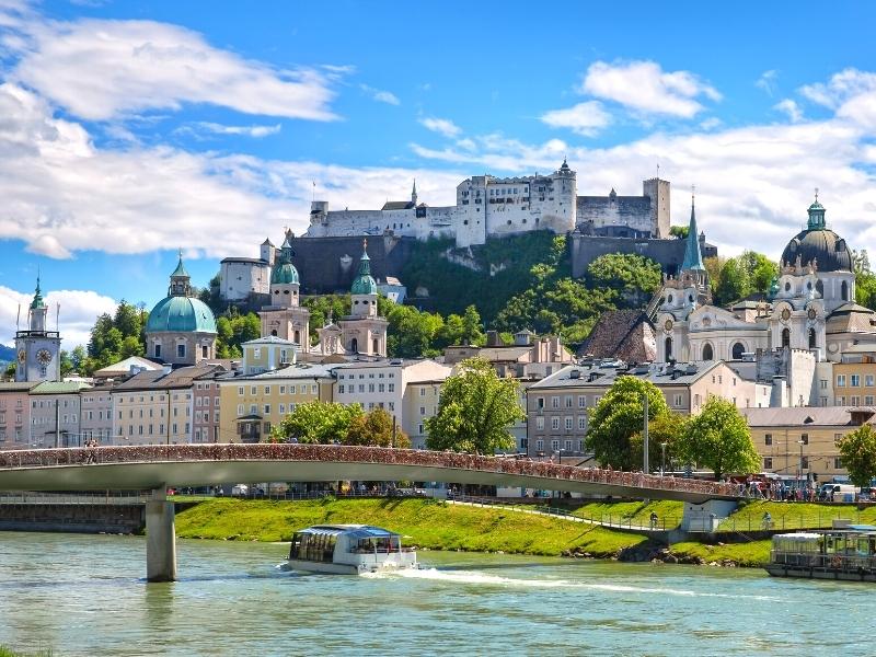 Hohensalzburg Fortress sits on a hill overlooking Salzburg, Austria with the Salzach River in the foreground spanned by a pedestrian bridge