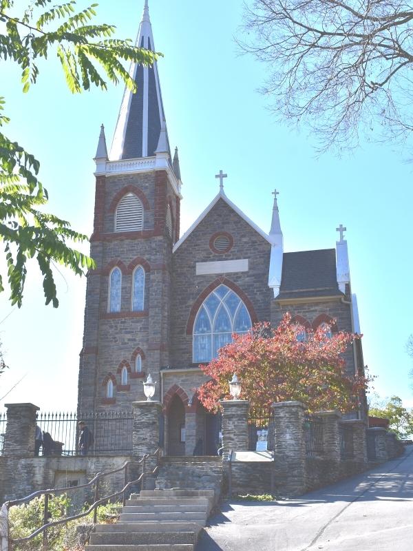 St Peter's Catholic church features red brick highlights around it's stained glass windows with a spire reaching to the blue sky, sits on the hillside above the Lower Town in Harpers Ferry, WV