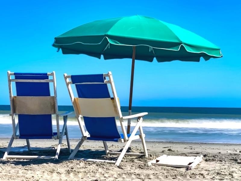 Blue beach chairs and a teal beach umbrella sit on the beach in front of the ocean