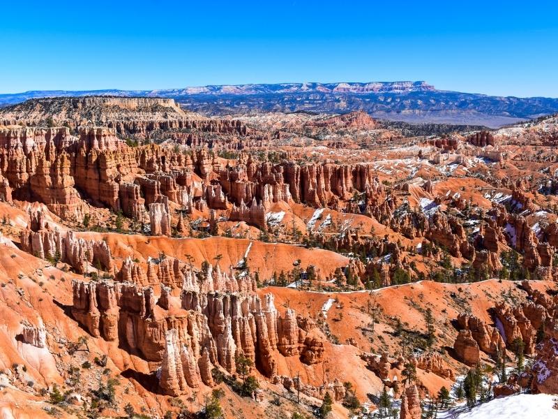 The bright orange rocks of the Bryce Amphitheater glow under a clear blue sky, with a few remaining snow piles that are slowly melting