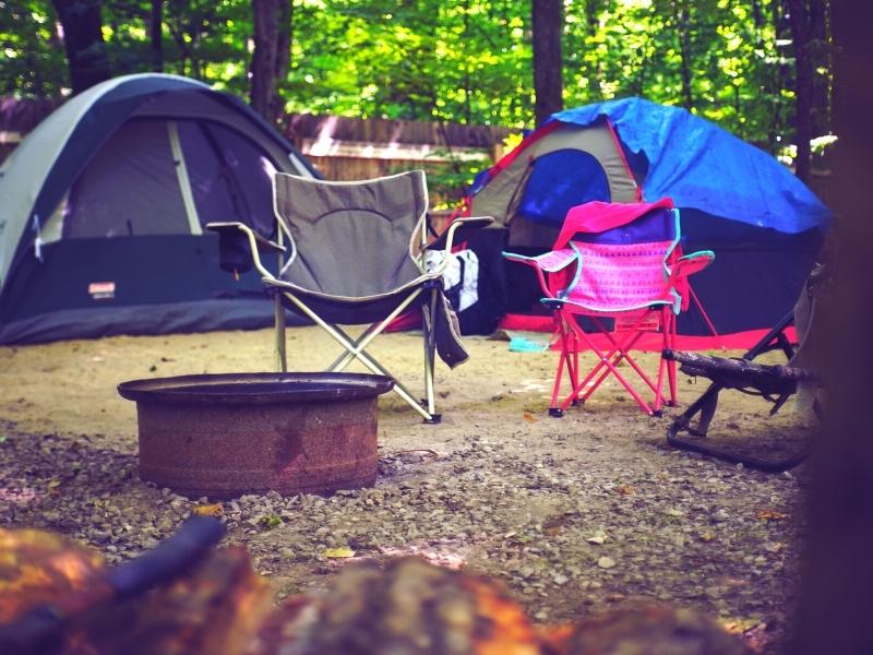 Tents and camp chairs are set up around a fire ring at a campsite in a forest