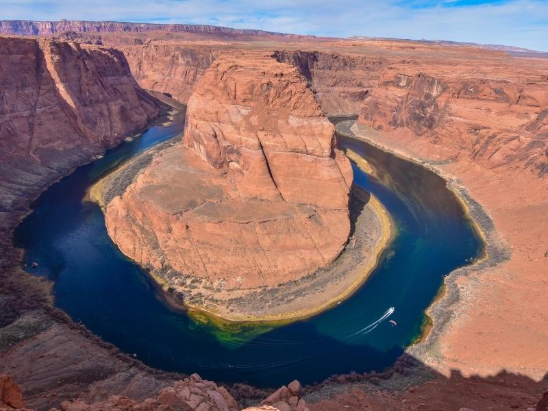 A boat cruises down the deep blue waters of the Colorado River, surrounded by the orange canyon walls at Horseshoe Bend, Arizona