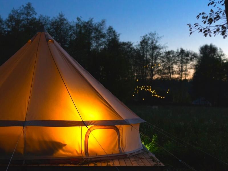 A canvas tent glows orange against the growing darkness as sun sets behind some trees