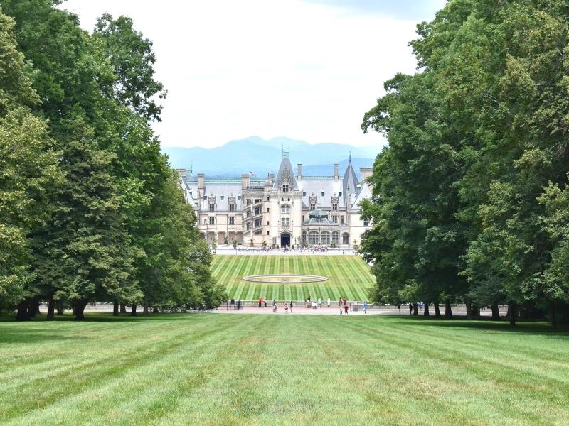 The Biltmore House sits below with Blue Ridge Mountains in the distance, the green striped Front Lawn, and the sloping green lawn flanked by large green trees