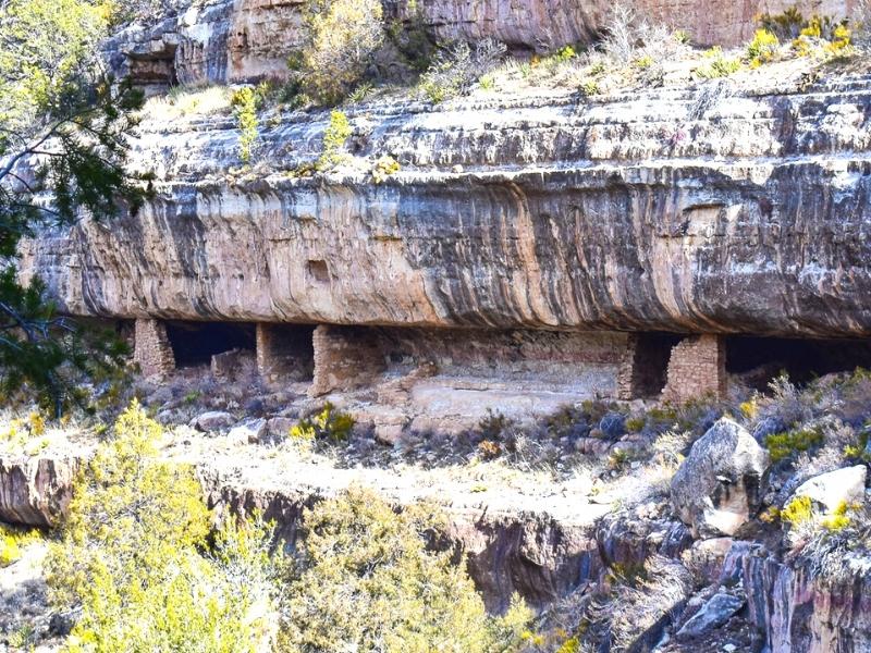 A tan rock cliff is stained black from years of rain but still shelters the remains of a Native American cliff dwelling set under the cliff ledge at Walnut Canyon National Monument