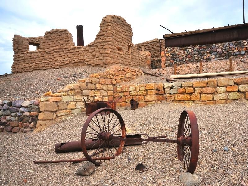 Historic ruins of the Harmony Borax Works in Death Valley National Park, with rusting leftovers of a wagon axle and stone buildings without roofs.