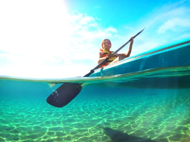 A woman in a yellow lifejacket kayaks through clear blue-green waters on a sunny day