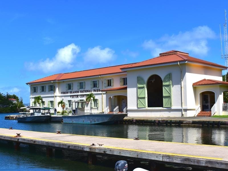 The Virgin Islands National Park Visitor Center is a white stucco building with red tin roof on the water in Cruz Bay, St John