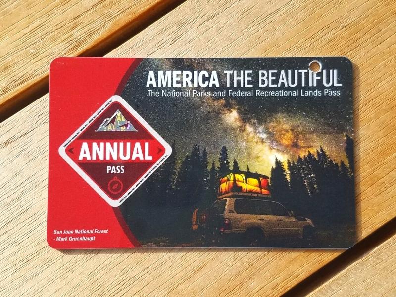 An America the Beautiful pass sits on a wooden table, with a red band on the left and a hole punch clearly visible through the photo of a starry night sky