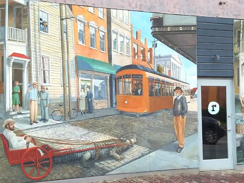 A mural depicting old town Charleston, SC with a cable car and a man in a rickshaw pulled by an alligator