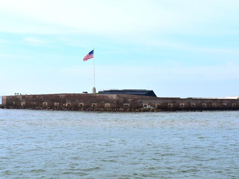 An American flag flies proudly over the brick walls of Fort Sumter, surrounded by Charleston Harbor waters under a blue and white sky